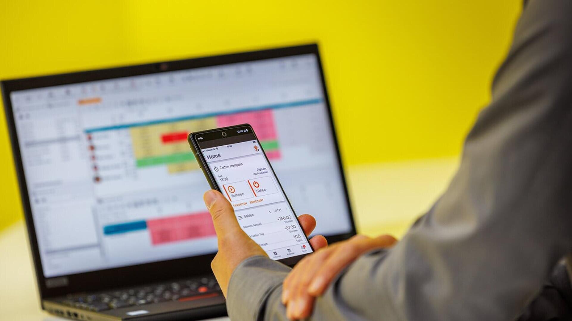 A person in a gray shirt holds a smartphone displaying a task management app, while a laptop with a color-coded spreadsheet is open in the background, all set against a yellow wall.