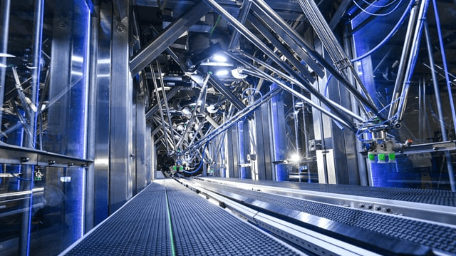 Industrial robotic arms operate inside a brightly lit, high-tech factory with blue lighting, metallic surfaces, and a mesh walkway running through the machinery.