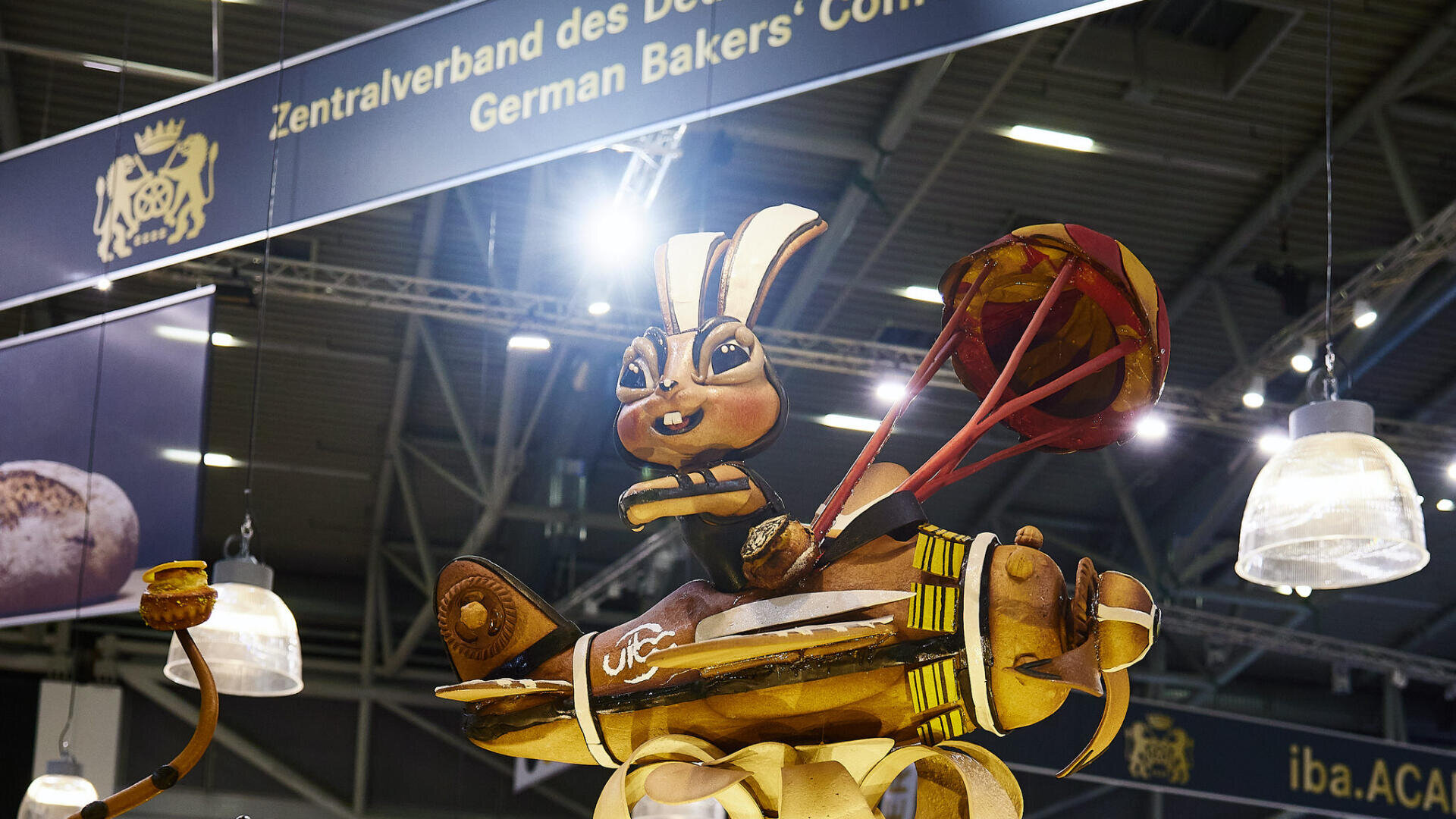Eine große, detaillierte Brotskulptur eines anthropomorphen Kaninchens, das eine Trommel und Trommelstöcke hält, wird auf einer deutschen Bäckertagung ausgestellt, wobei im Hintergrund Banner hängen.