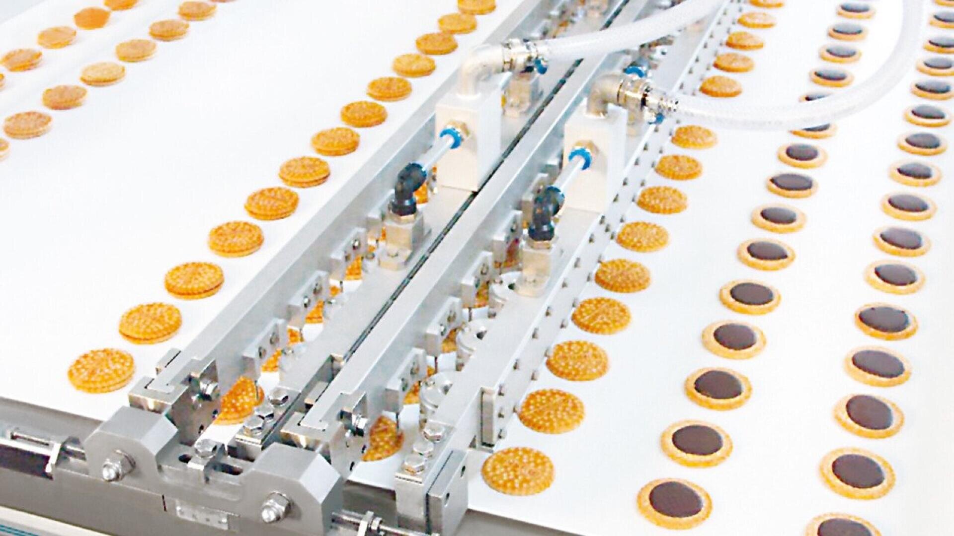 Rows of cookies are being processed on a conveyor belt by an automated machine with metallic components and tubes, likely as part of a food production line in a factory.