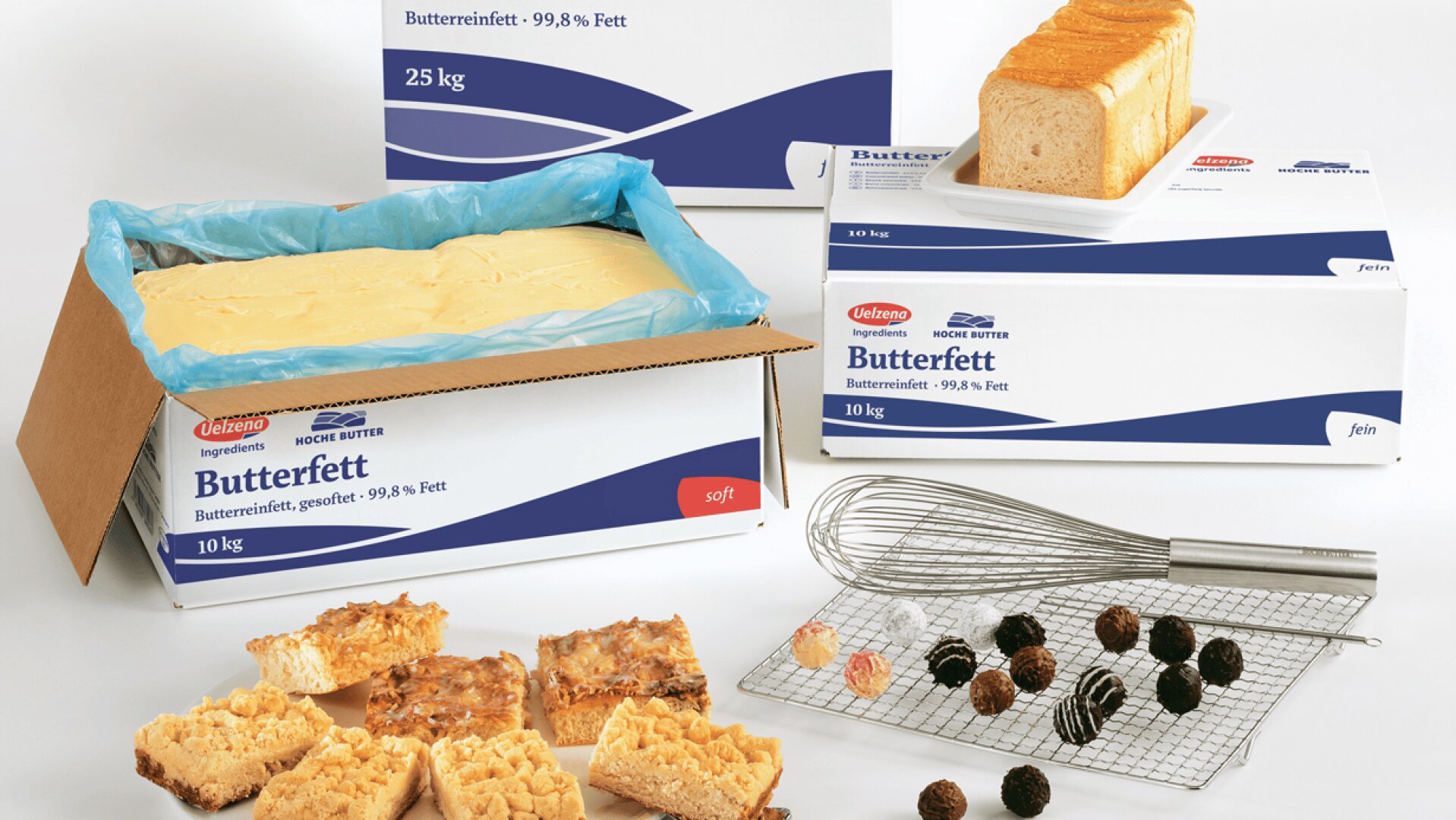 Schachteln mit der Aufschrift Butterfett sind mit einem großen offenen Behälter mit Butterfett, einem Laib Weißbrot, verschiedenen Backwaren, Pralinen und einem Schneebesen auf einem Kühlgestell auf einer weißen Fläche abgebildet.