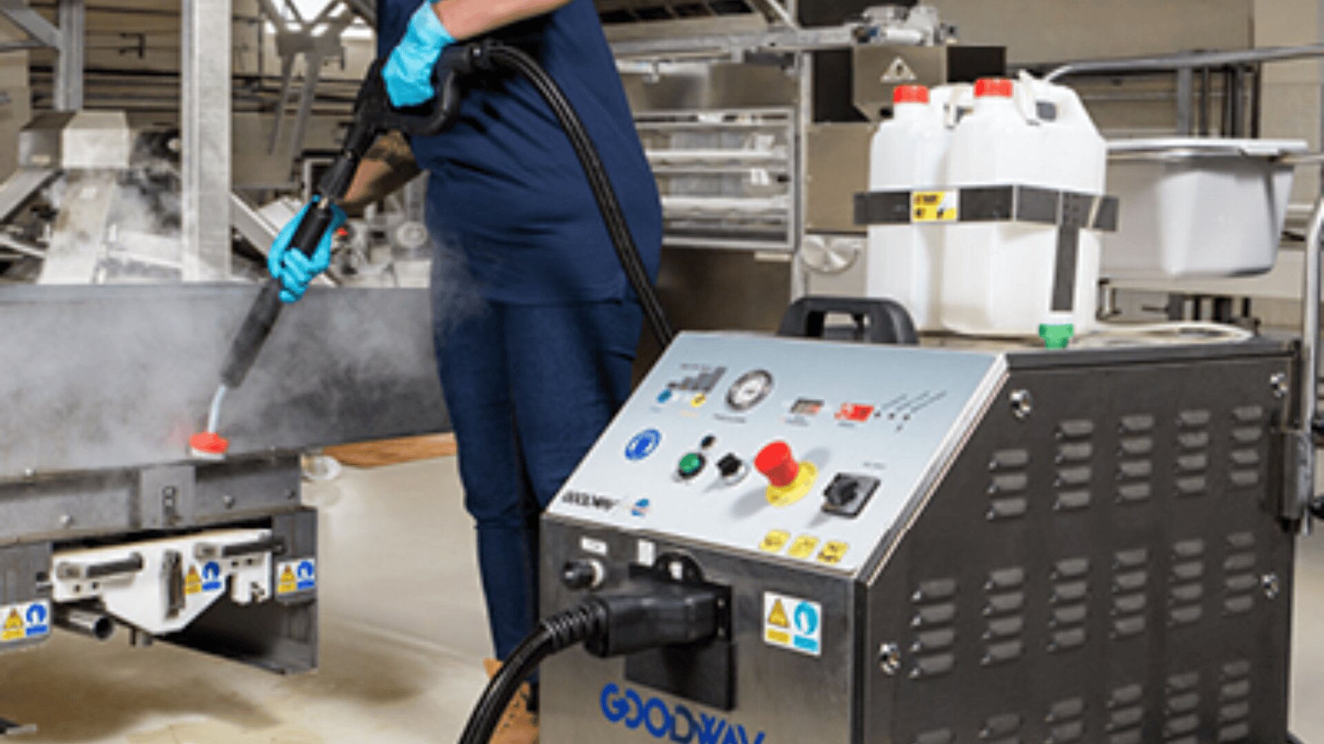 A worker wearing gloves uses a steam cleaning machine labeled Goodway to clean industrial equipment, with plastic chemical containers placed nearby.