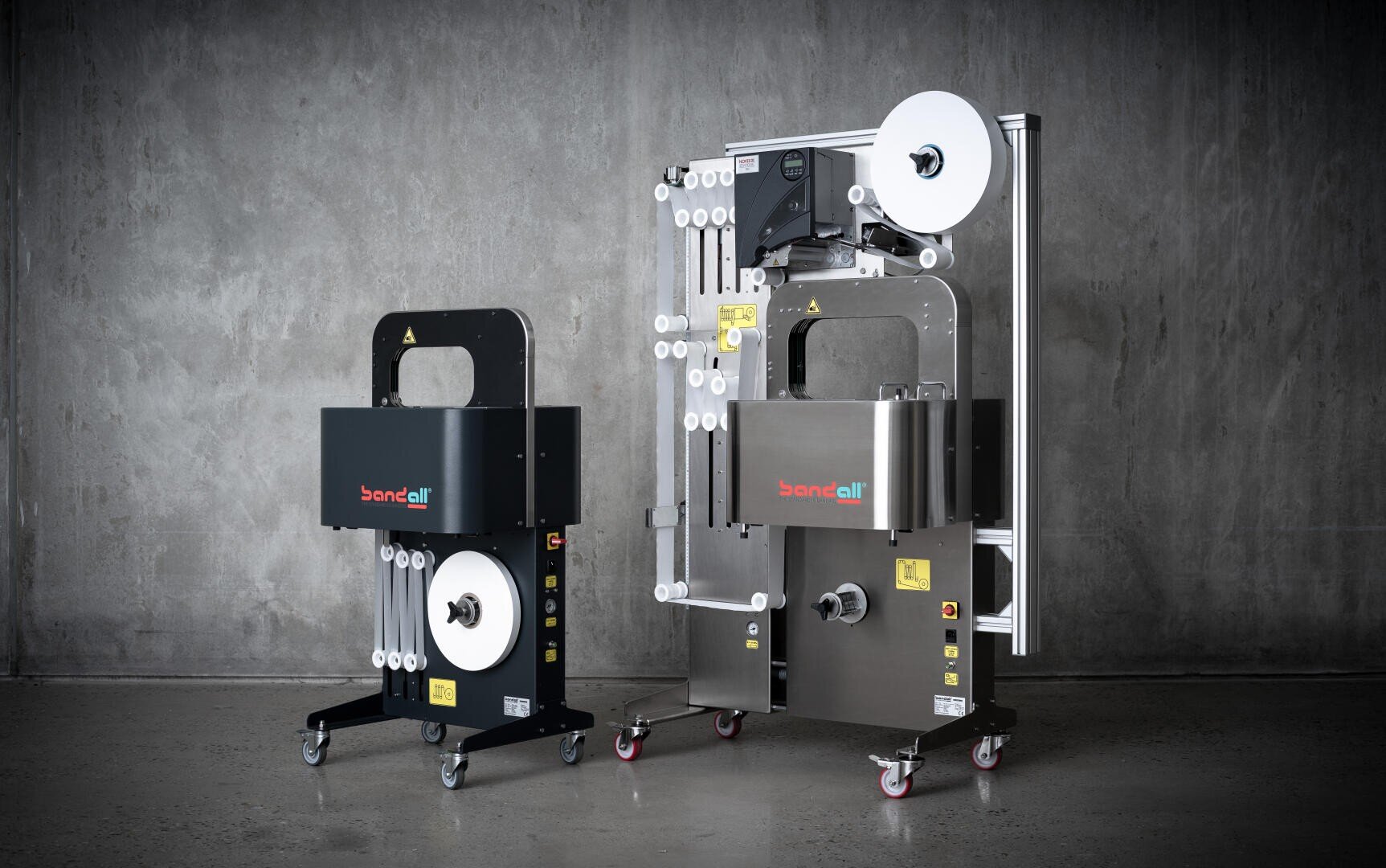 Two industrial banding machines with wheels stand side by side against a gray concrete wall. The machines have large rolls of banding material and display the bandall logo on their fronts.