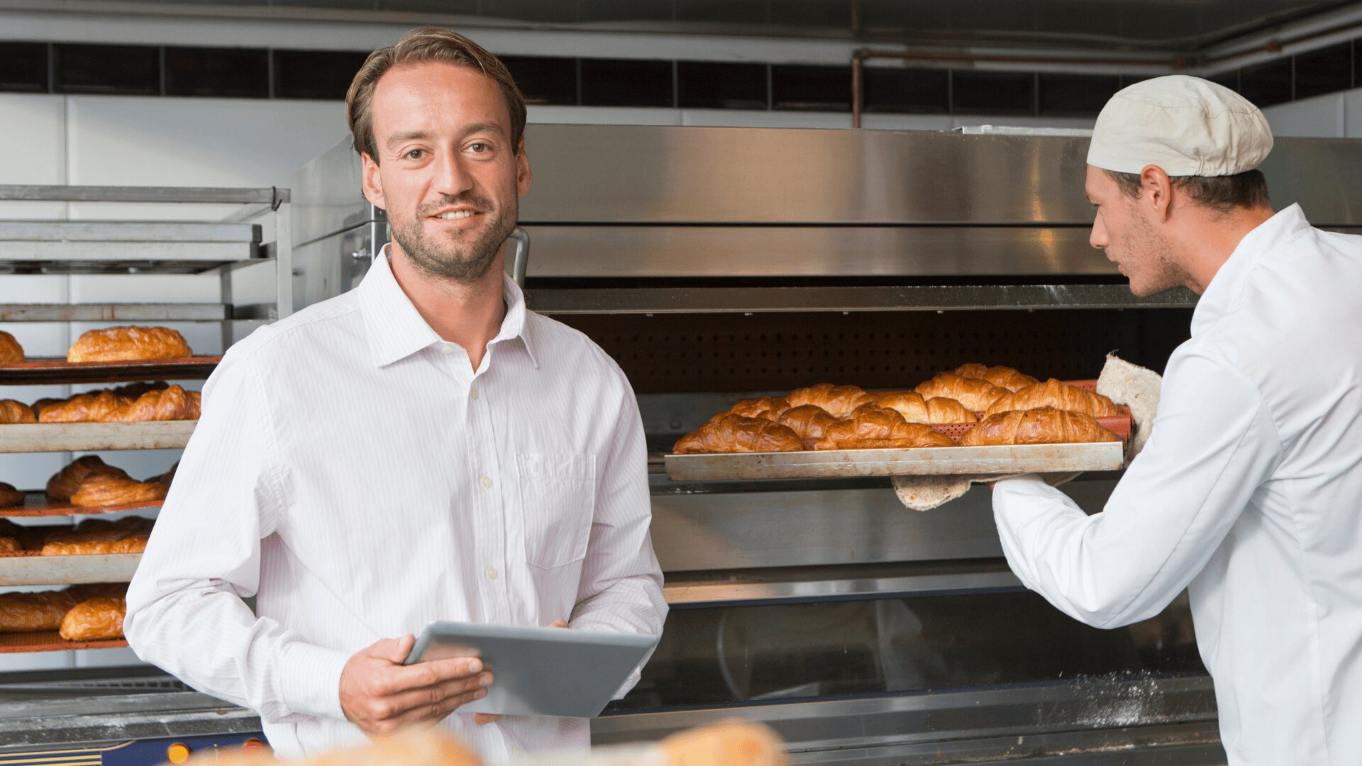 Ein Mann in einem weißen Hemd hält ein Tablet in der Hand und lächelt in die Kamera, während eine andere Person in einer Bäckeruniform ein Tablett mit Croissants aus einem Industrieofen in einer Bäckerei nimmt.