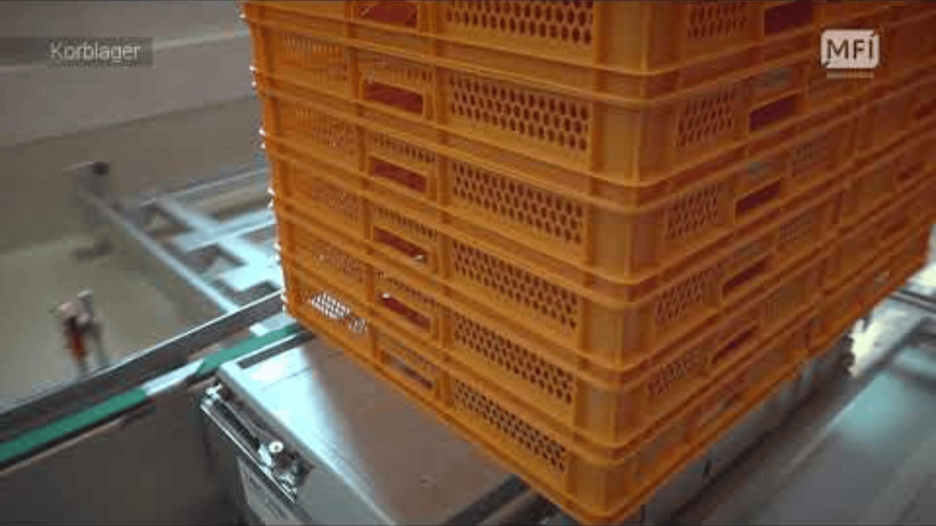 Ein Stapel orangefarbener Kunststoffkisten befindet sich auf einem Metallförderband in einer Industrieumgebung. Die Kisten haben Lüftungslöcher, und die Maschinen scheinen Teil eines automatisierten Prozesses zu sein.