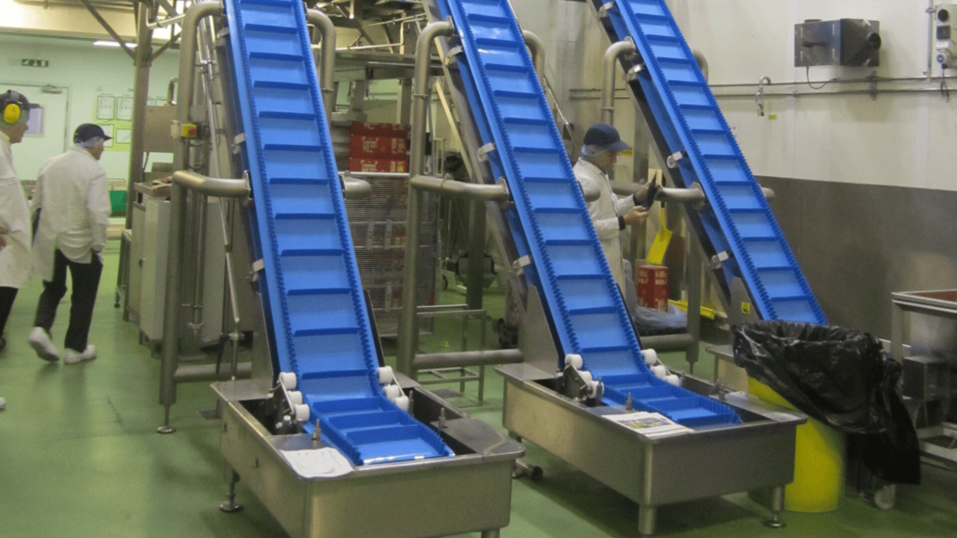 Three blue conveyor belts in a food processing facility with workers in white coats, hairnets, and masks; stainless steel equipment and bins visible in the clean, organized workspace.