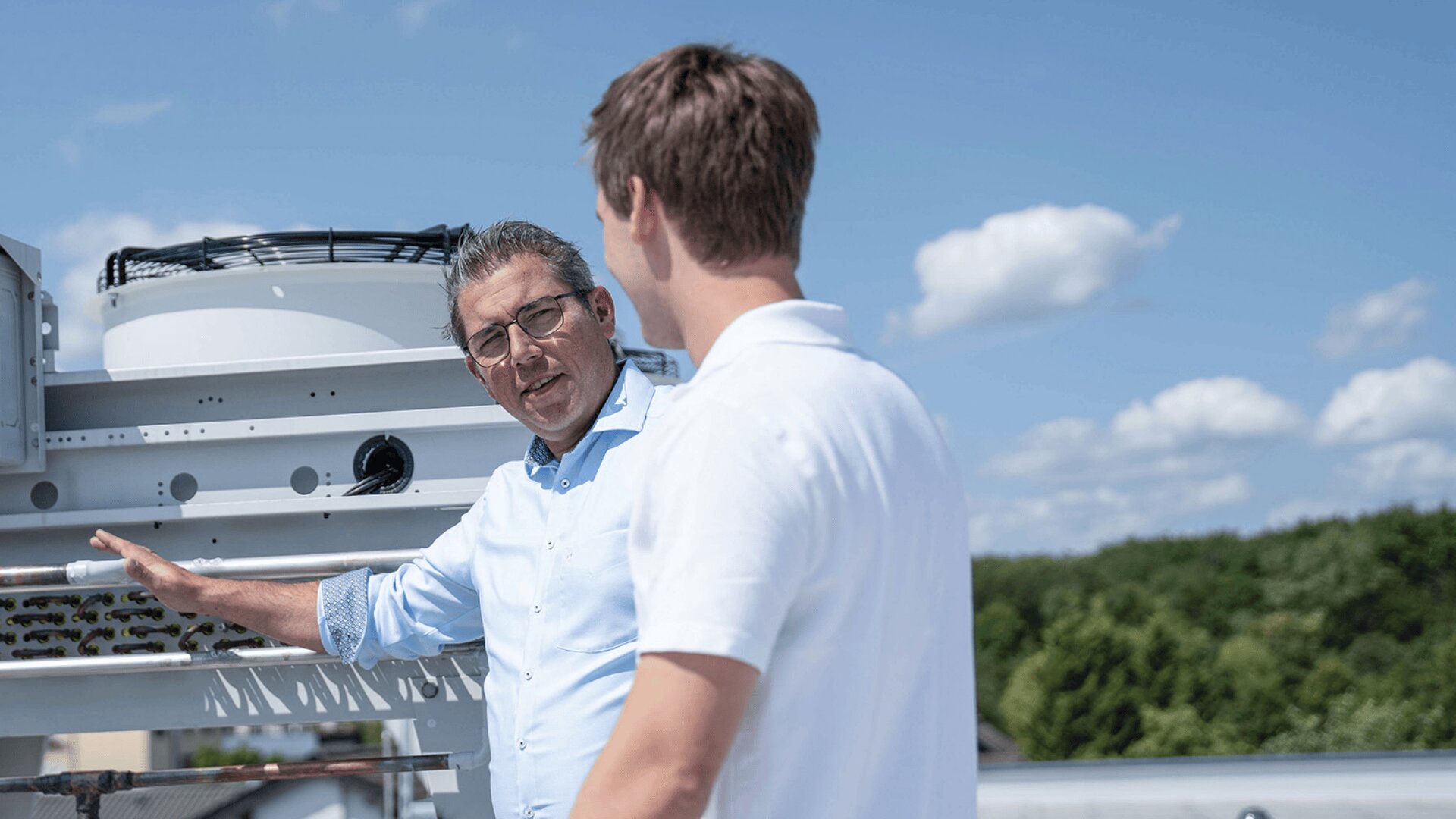 Two men in white shirts are talking outdoors near industrial equipment on a rooftop, with a blue sky and green trees in the background.