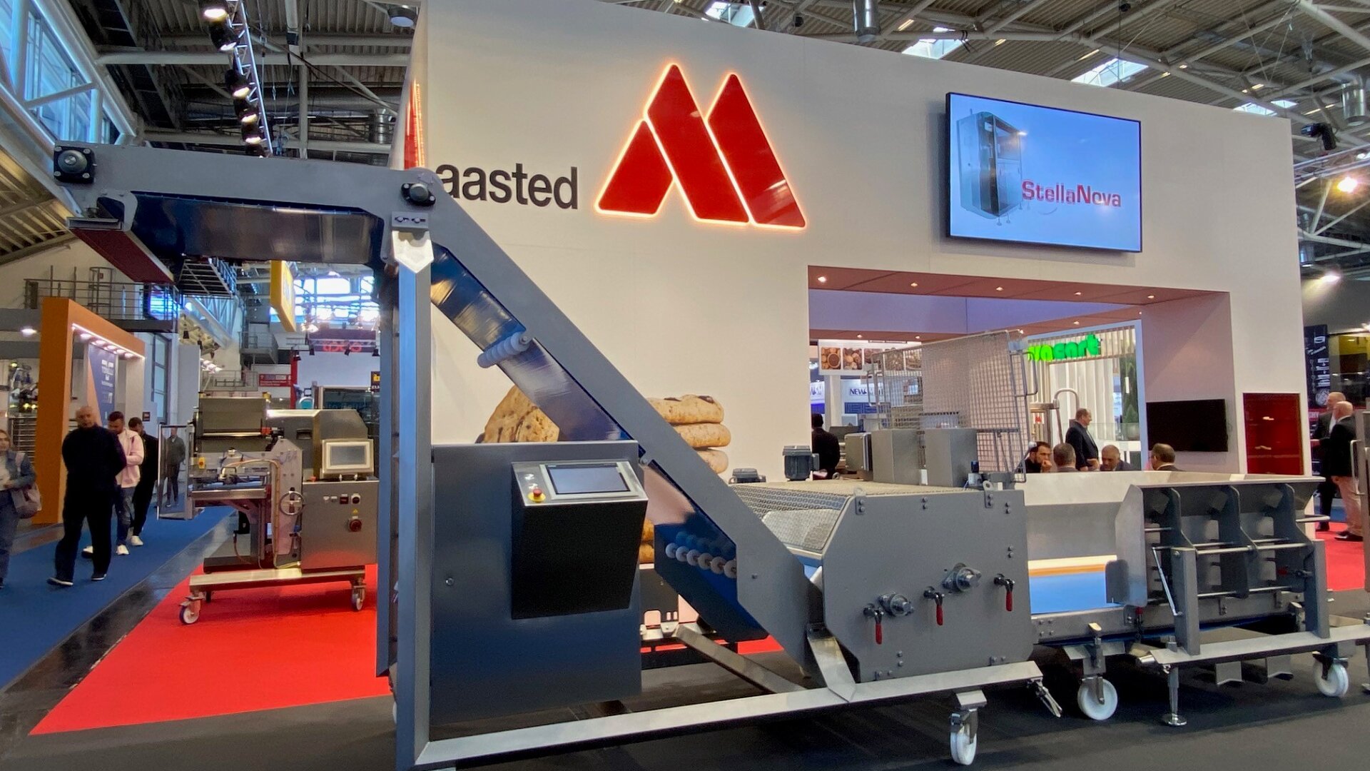 A large industrial machine is displayed at a trade show booth with a sign reading maasted and a glowing red logo. People and other booths are visible in the background. A screen displays StellaNova.