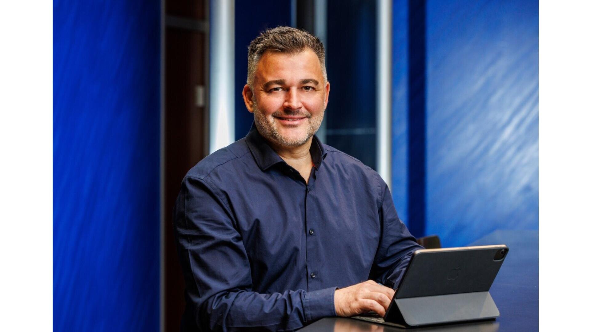 A man with short gray hair and a beard, wearing a dark blue shirt, sits at a desk with a tablet in front of him, smiling at the camera. The background features blue walls and glass panels.