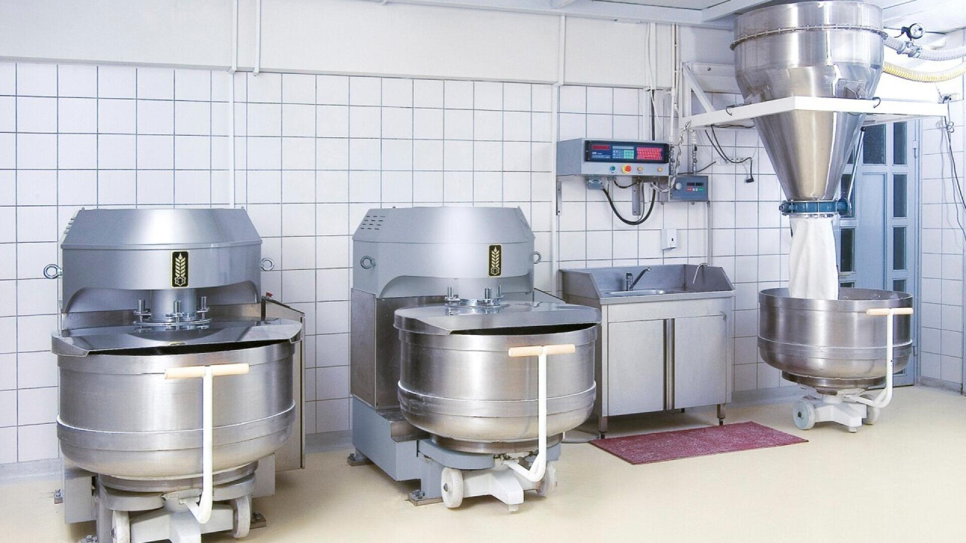 Industrial bakery kitchen with three large stainless steel dough mixers, a metal sink, wall-mounted controls, and a flour dispenser feeding into one of the mixers. The room has tiled walls and a clean, organized appearance.