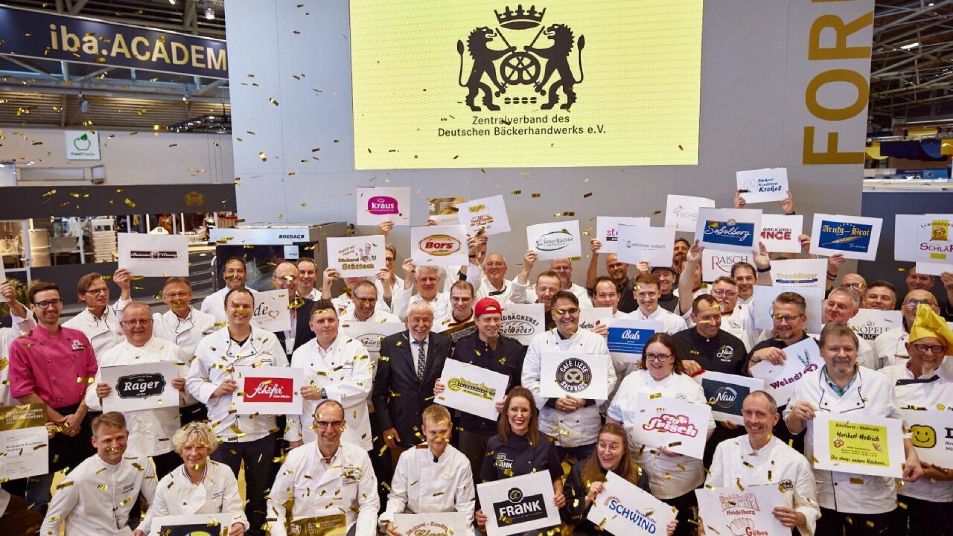 A large group of people in chef uniforms hold up bakery brand signs, posing and smiling under falling confetti at an indoor event. A yellow screen behind them shows the logo for the German Bakers’ Association.