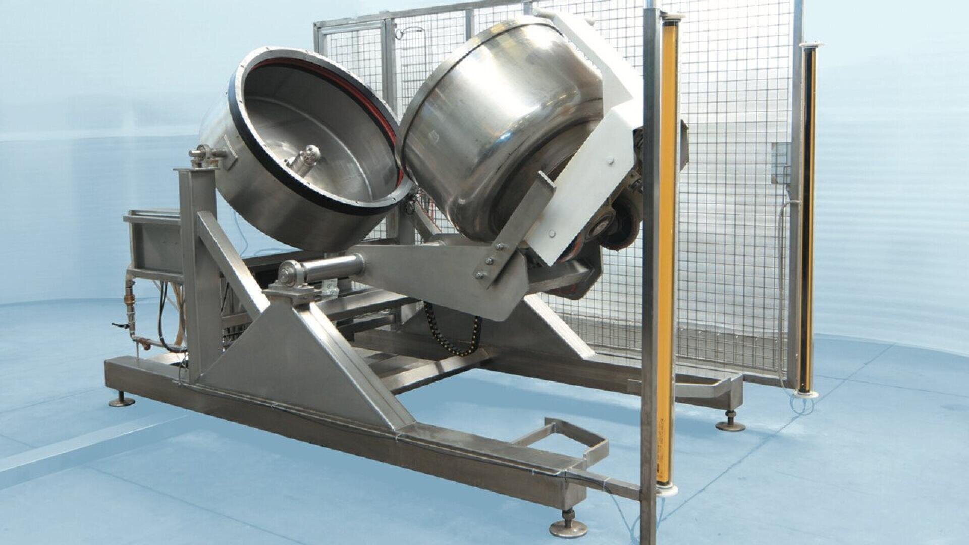 A large industrial stainless steel mixer with twin rotating drums is set in a clean, blue-walled room and protected by a wire safety guard. The equipment is used for mixing or blending food or chemical products.