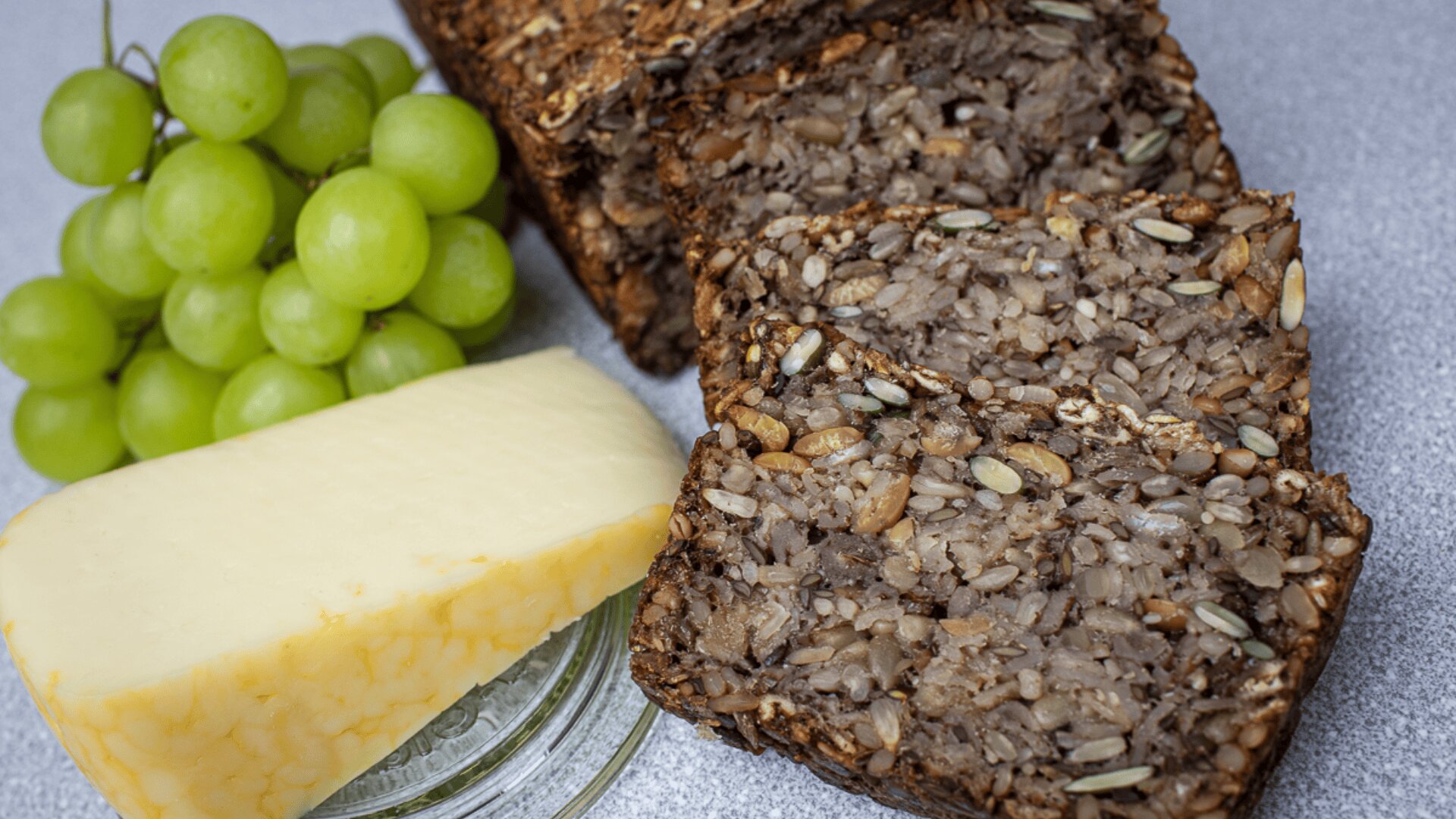 A cluster of green grapes, a wedge of cheese, and several slices of seed-filled multigrain bread are arranged on a light gray surface.