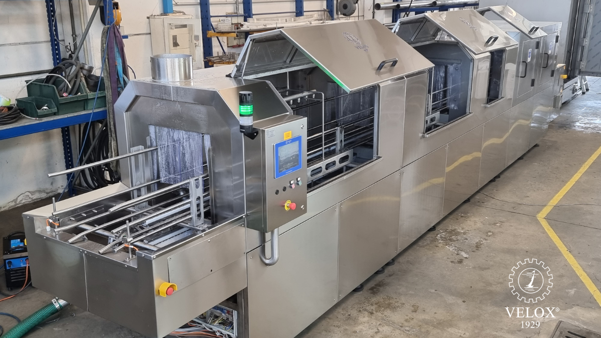 A large stainless steel industrial machine with control panels and open compartments, situated in a workshop. The brand VELOX 1929 logo is visible in the lower right corner of the image.