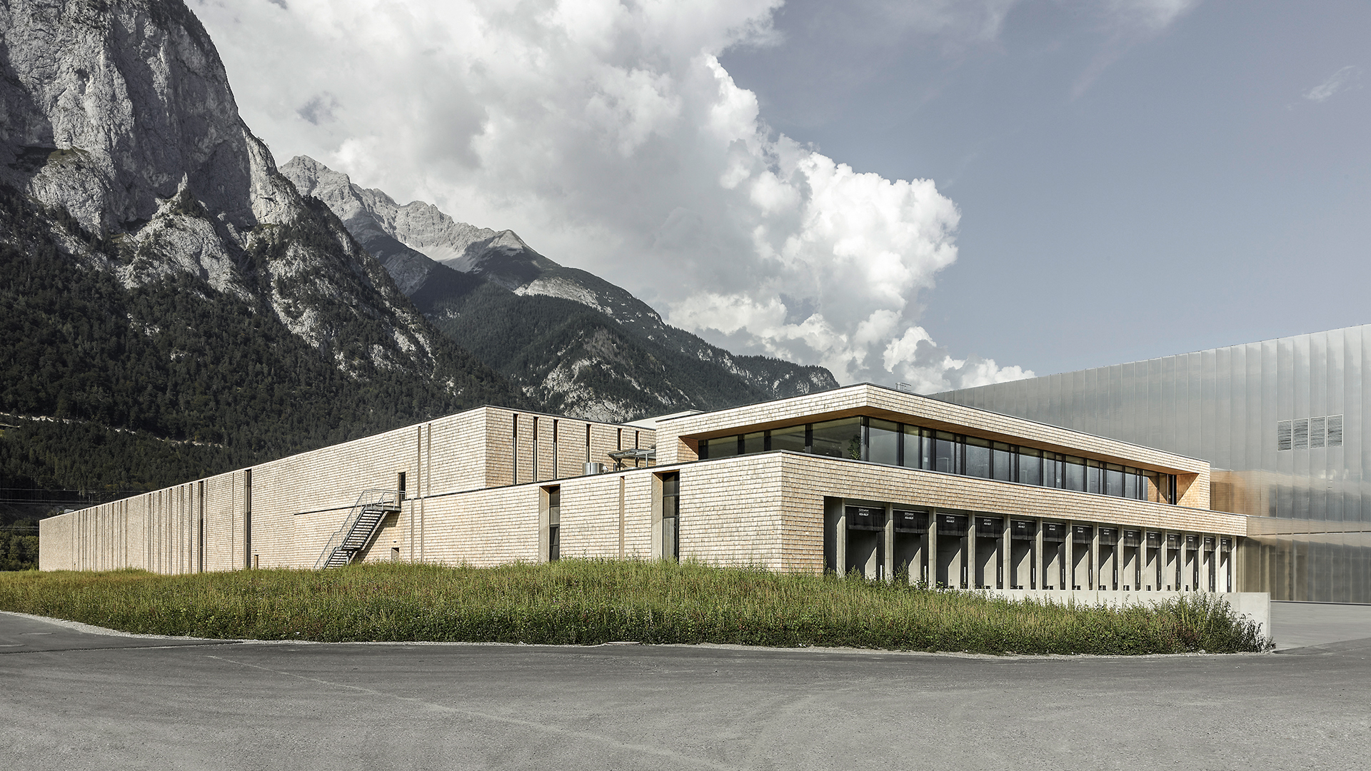 Ein modernes, niedriges Gebäude mit großen Fenstern und hellen Backsteinmauern steht in der Nähe einer Rasenfläche, im Hintergrund hohe Berge unter einem teilweise bewölkten Himmel.