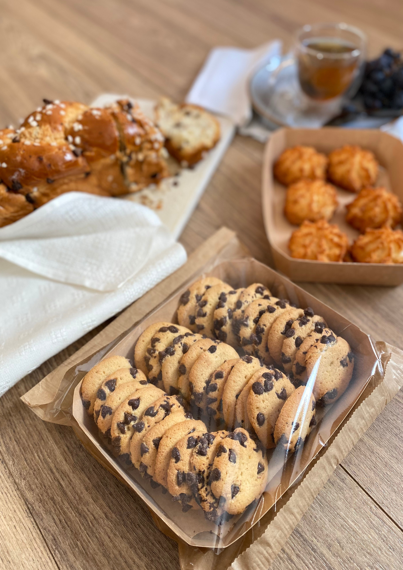 Im Vordergrund ein Tablett mit Chocolate Chip Cookies, im Hintergrund Kokosmakronen, ein geflochtener Brotlaib, eine Tasse Tee und Weintrauben auf einem Holztisch.