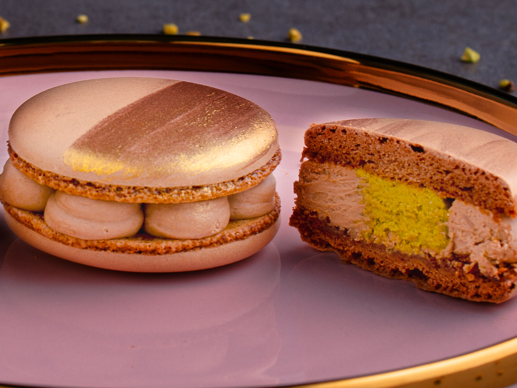 Two elegant macarons rest on a pink plate with a gold rim. One macaron is whole with gold decoration, while the other is cut in half, revealing creamy light brown and yellow fillings.
