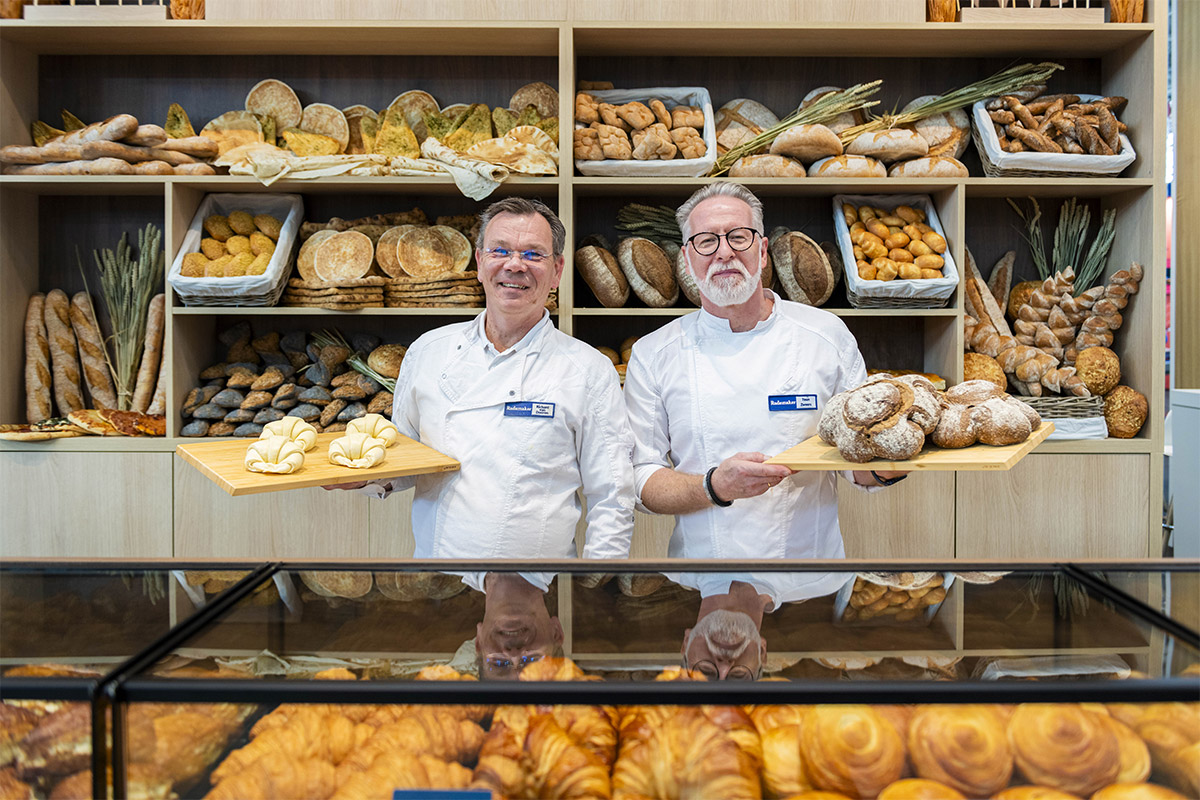Zwei Bäcker in weißen Kitteln stehen in einer Bäckerei und halten jeweils ein Tablett mit Brot in den Händen. Hinter ihnen befinden sich Regale mit verschiedenen Brot- und Brötchensorten. Vorne in einer Glasvitrine werden Gebäckstücke präsentiert.