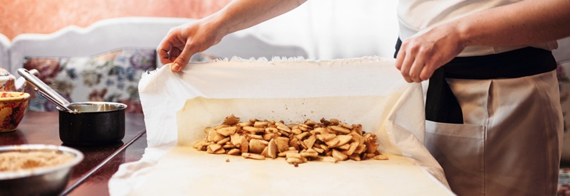 Eine Person bereitet ein Gebäck zu, indem sie eine Füllung aus Apfelscheiben auf eine Teigplatte streicht, die zum Backen aufgerollt werden soll. Die Szene spielt in einer gemütlichen Küche.