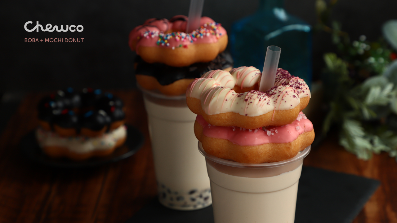 Two cups of bubble tea are topped with colorful mochi donuts; one donut is pink with sprinkles, the other white with pink flakes. Another donut with chocolate icing and sprinkles can be seen out of focus in the background.