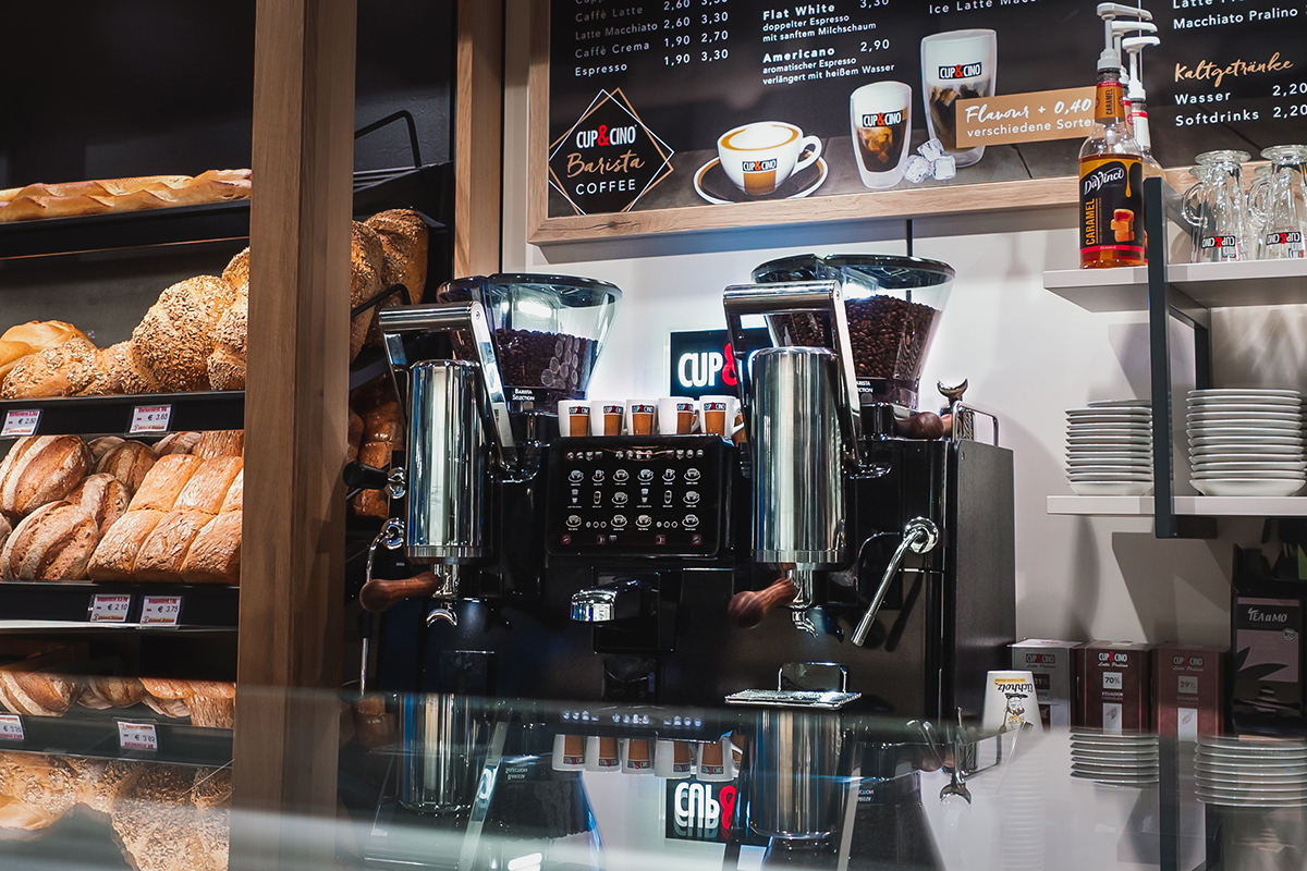 A coffee machine with two grinders sits on a counter in a bakery, surrounded by loaves of bread on shelves and menu boards above. Small glass cups and packaged goods are also visible.
