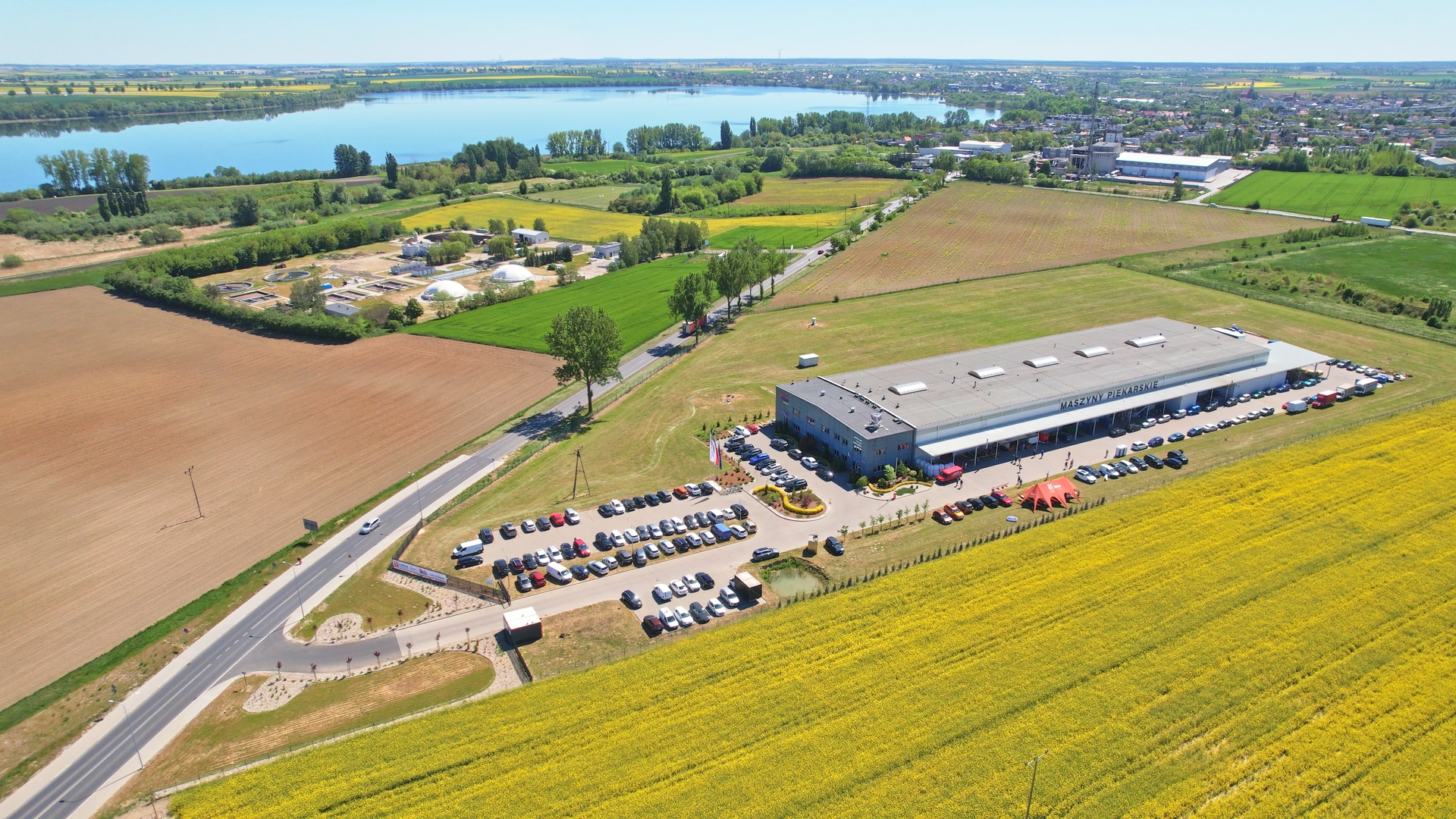 Aerial view of a modern building with a parking lot filled with cars, surrounded by yellow fields, farmland, a road, and a river in the background, with a town visible in the distance.