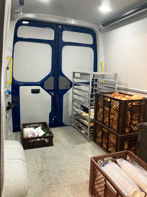 Inside view of a delivery van with stacked metal racks holding bread and pastries, and brown crates filled with packaged food items and cups on the floor near the closed rear doors.