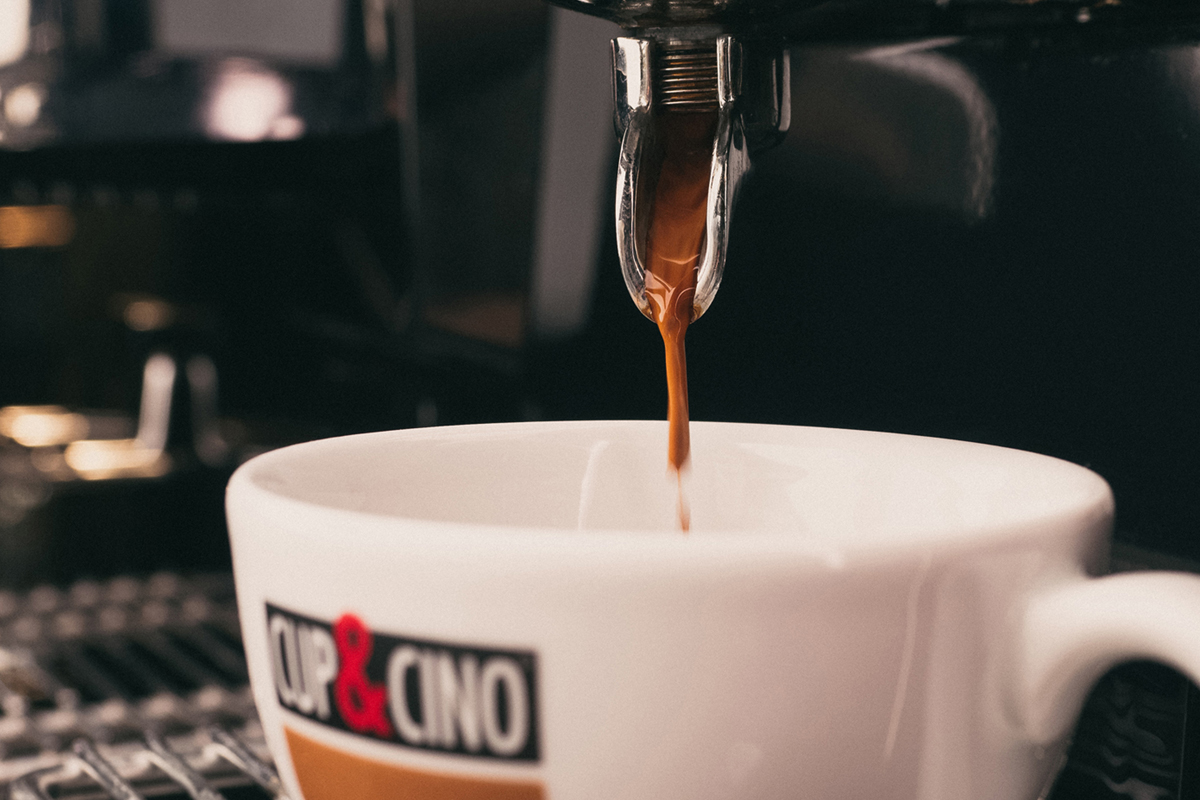Close-up of espresso machine pouring coffee into a white cup with a CUP & CINO logo. The coffee stream is rich and brown, and the scene has a warm, inviting atmosphere.
