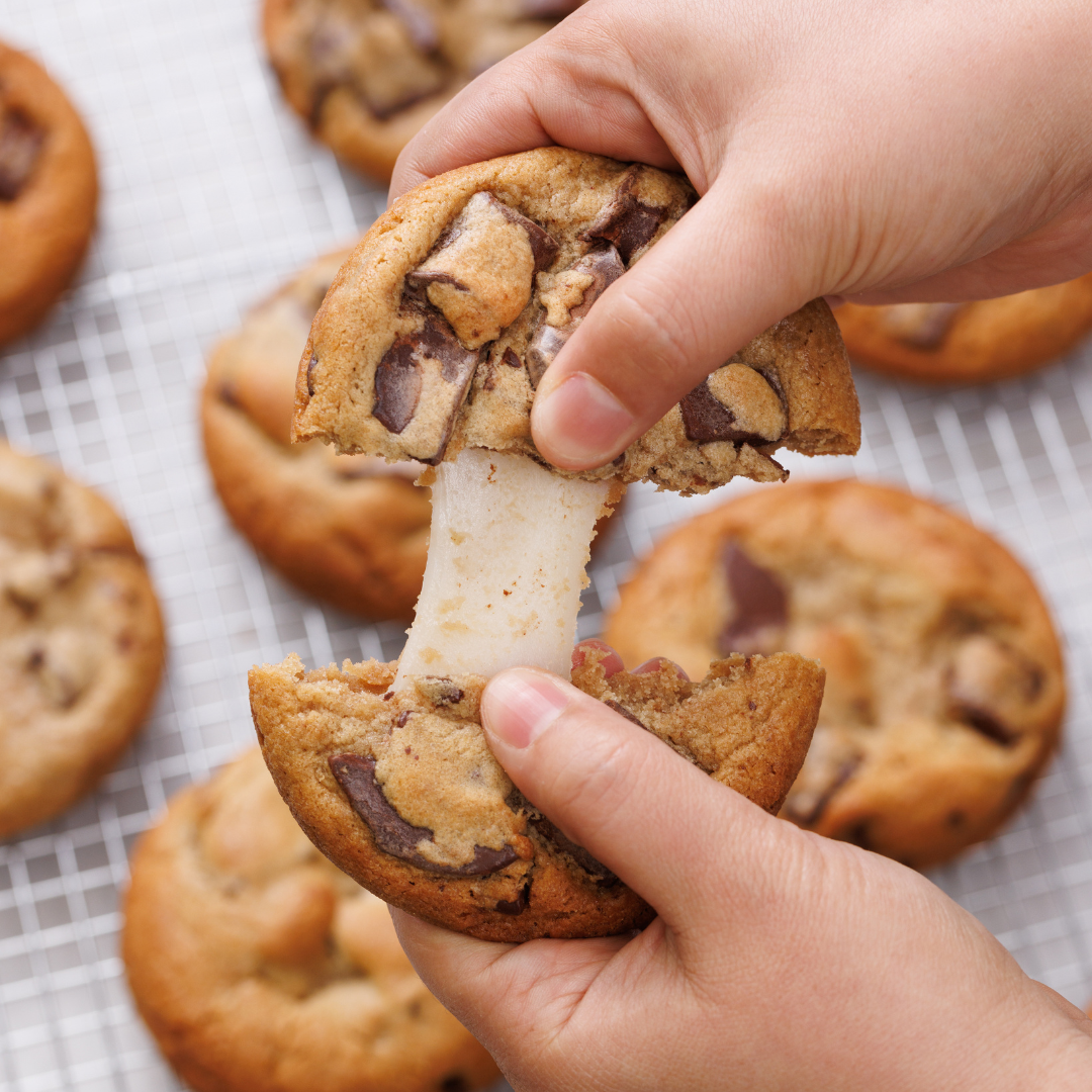 Two hands cut up a freshly baked chocolate cookie with a sticky, melted center. In the background you can see more cookies on a cake rack.
