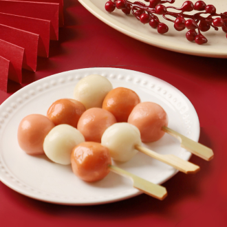 Ein weißer Teller mit Spießen aus runden, rosa-weißen Dango (japanische Reisklöße) auf rotem Untergrund. Im Hintergrund sind dekoratives rotes Papier und ein Teller mit roten Beeren zu sehen.
