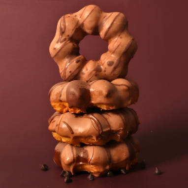 Vier schokoladenglasierte Donuts sind vertikal vor einem braunen Hintergrund gestapelt, unten sind Schokoladenstückchen verstreut. Der oberste Donut steht aufrecht und sorgt für eine optisch ansprechende Präsentation.