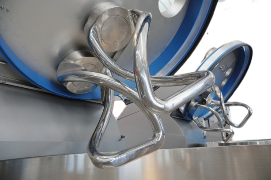 Nahaufnahme großer, glänzender industrieller Knethaken, die an einer kommerziellen Knetmaschine angebracht sind, mit metallischen Oberflächen und blauen Akzenten im Hintergrund.