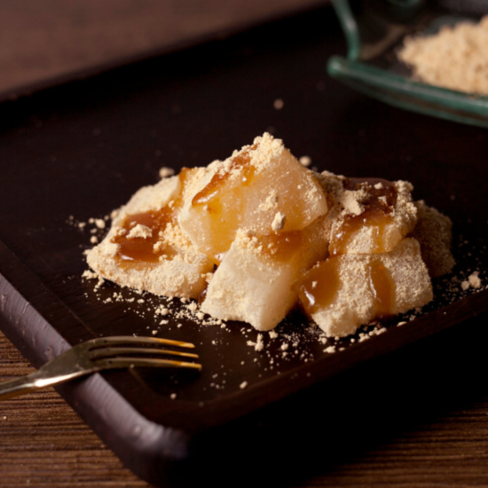 Three pieces of transparent mochi are sprinkled with brown syrup and kinako powder and served on a dark wooden tray. Next to it is a small fork. In the background is a small bowl with additional powder.