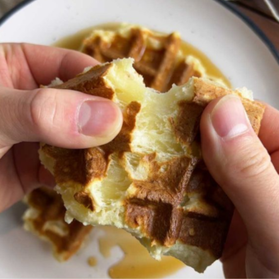 Two hands break a waffle over a white plate of syrup.