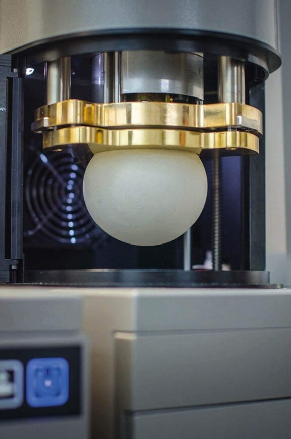 Eine glatte, runde Teigkugel wird in einer Metallmaschine von einer goldfarbenen Platte gepresst, wahrscheinlich zum Testen oder Formen. Im Hintergrund ist ein schwarzer Fächer zu sehen.