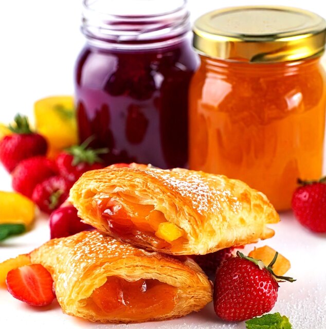 Zwei mit Früchten gefüllte Gebäckstücke sind vor Gläsern mit roter und orangefarbener Marmelade gestapelt, umgeben von frischen Erdbeeren und einigen gelben Fruchtstücken auf einer weißen Fläche. Das Gebäck ist mit Puderzucker bestäubt.