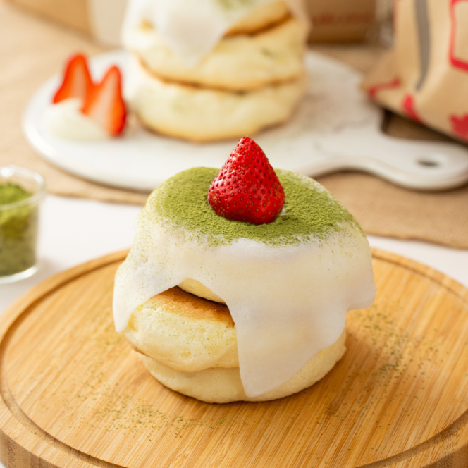 A fluffy Japanese pancake with white icing, matcha powder and a whole strawberry, served on a wooden plate with more pancakes and sliced strawberries in the background.