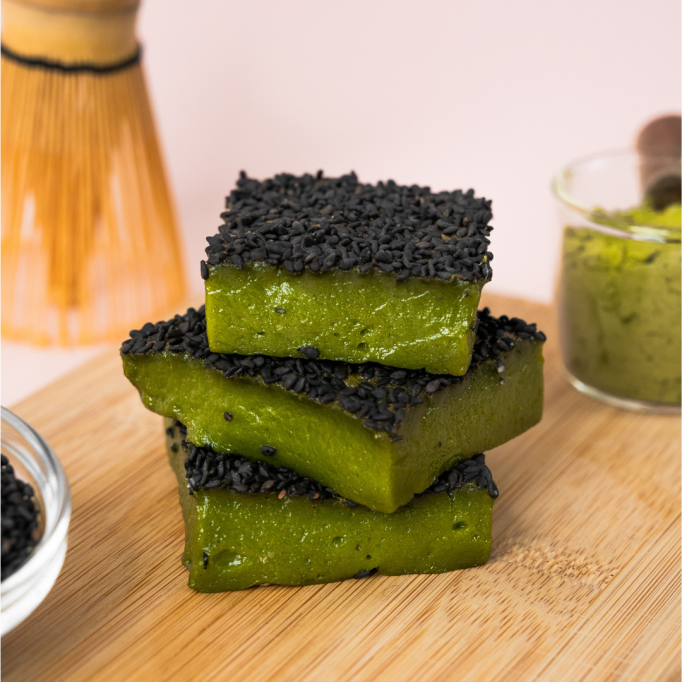 Three slices of green matcha cake topped with black sesame seeds are stacked on a wooden board, with matcha powder and utensils in the background.