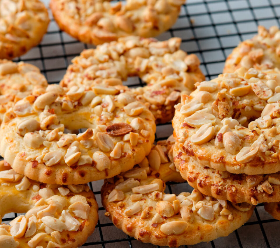 A close-up of round cookies topped with chopped peanuts, stacked on a black wire cooling rack. The cookies are golden brown and textured, with visible peanut pieces.
