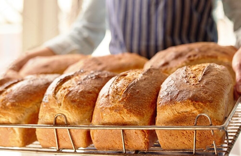 A person wearing a striped apron holds a cooling rack filled with several freshly baked loaves of bread, golden brown and crusty, in a bright kitchen setting.