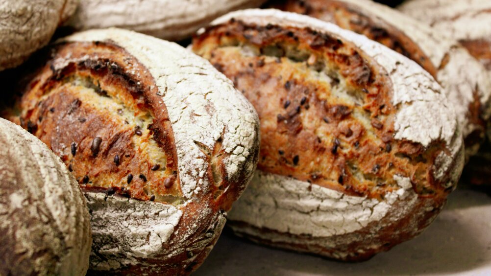 Several rustic loaves of bread with a crusty, browned exterior and a dusting of flour are stacked closely together. The loaves have a split top and visible seeds baked into the crust.