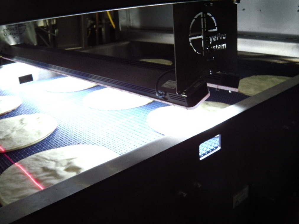 Runde Fladenbrote werden auf einem Förderband unter einer Maschine mit einem Laserscansystem hindurch bewegt, wahrscheinlich zur Qualitätskontrolle oder Inspektion in einer Lebensmittelverarbeitungsanlage.