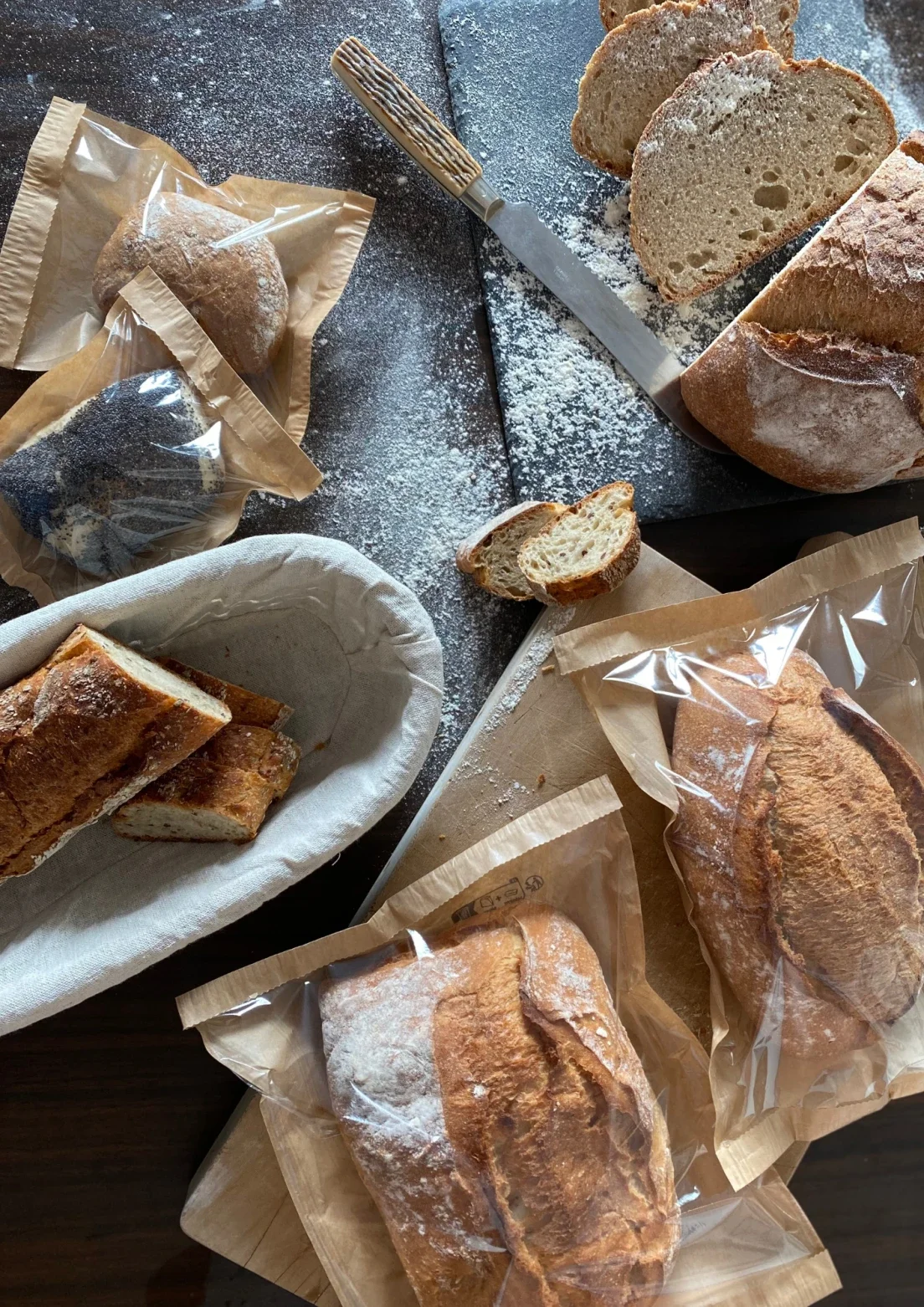 Ein Tisch mit verschiedenen Brotlaiben, teils geschnitten, teils ganz, in Papiertüten. Auf einem Schneidebrett liegen ein Brotmesser, etwas Mehl und ein paar Brotstücke.