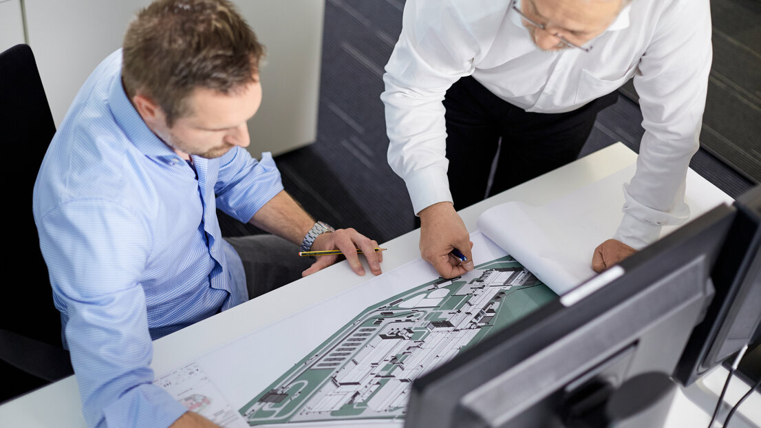 Zwei Männer in Geschäftskleidung prüfen an einem Schreibtisch Architekturpläne. Einer sitzt, der andere steht und beide konzentrieren sich auf die Zeichnungen vor einem Computermonitor.