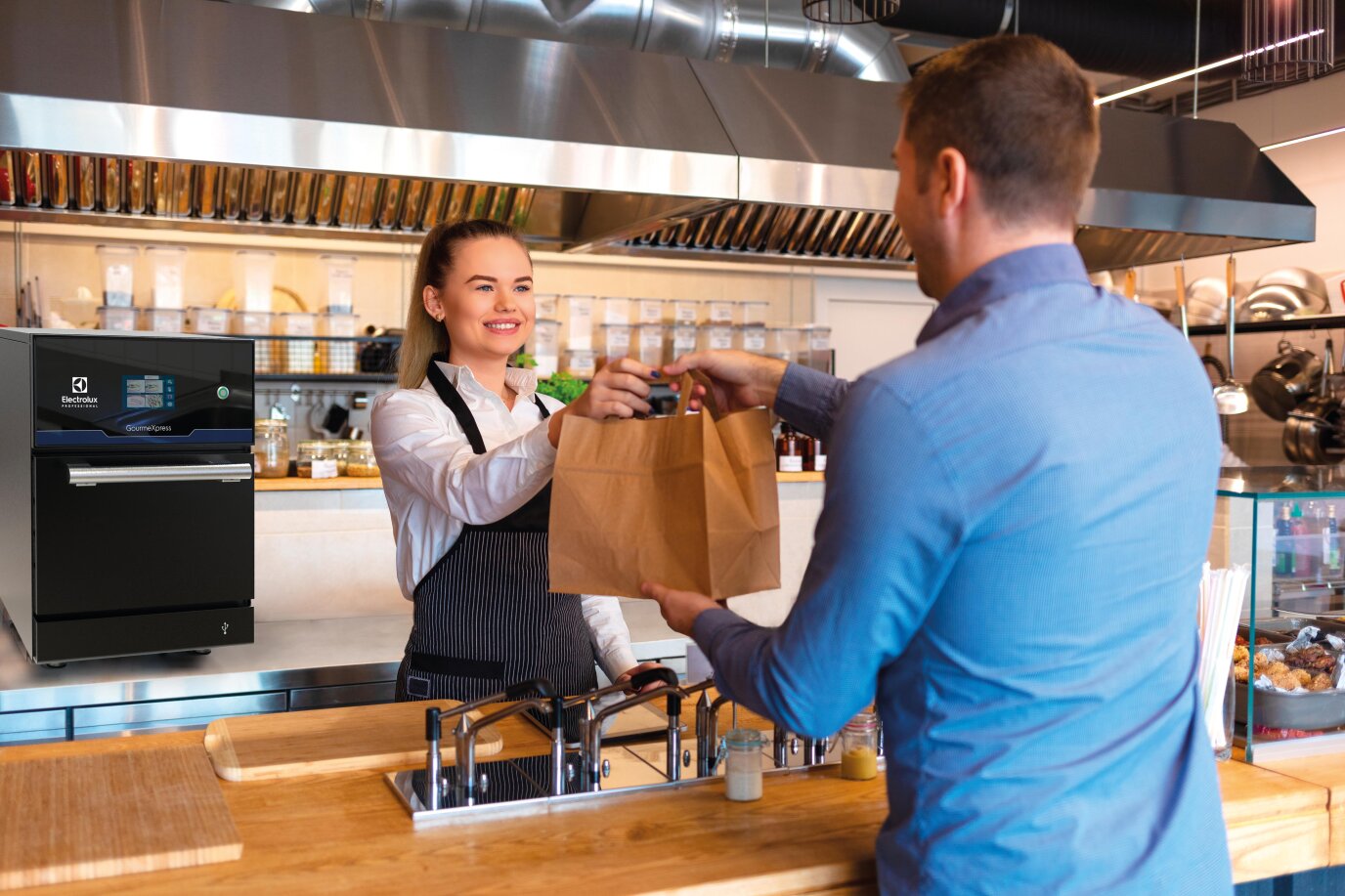 Eine lächelnde Frau in einer schwarzen Schürze reicht einem Mann an der Theke eines Restaurants eine Tüte zum Mitnehmen. Hinter ihr ist die Küche zu sehen, mit Kochgeräten und Regalen mit Zutaten.