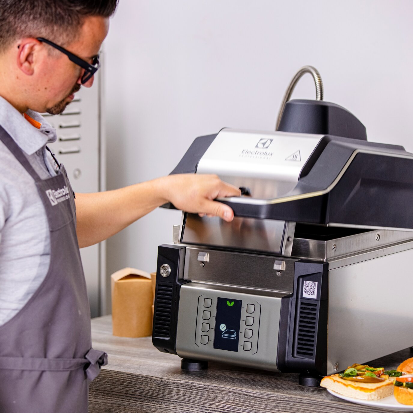 A person in glasses and an apron opens a modern countertop pizza oven, with a partially visible pizza on a plate nearby.