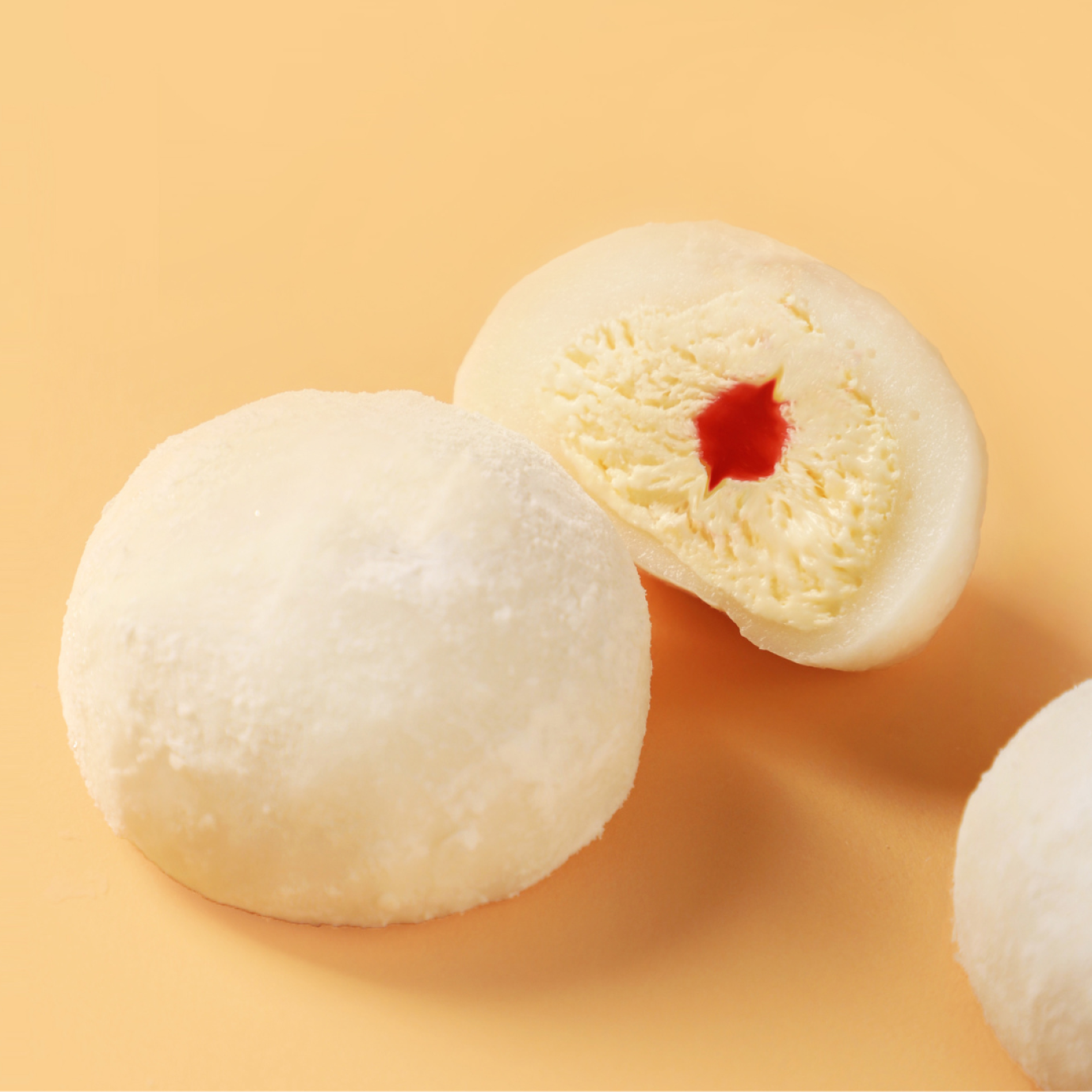 Two mochi ice cream scoops on a light peach background, one whole and one cut open to reveal creamy vanilla ice cream with a red fruit filling.