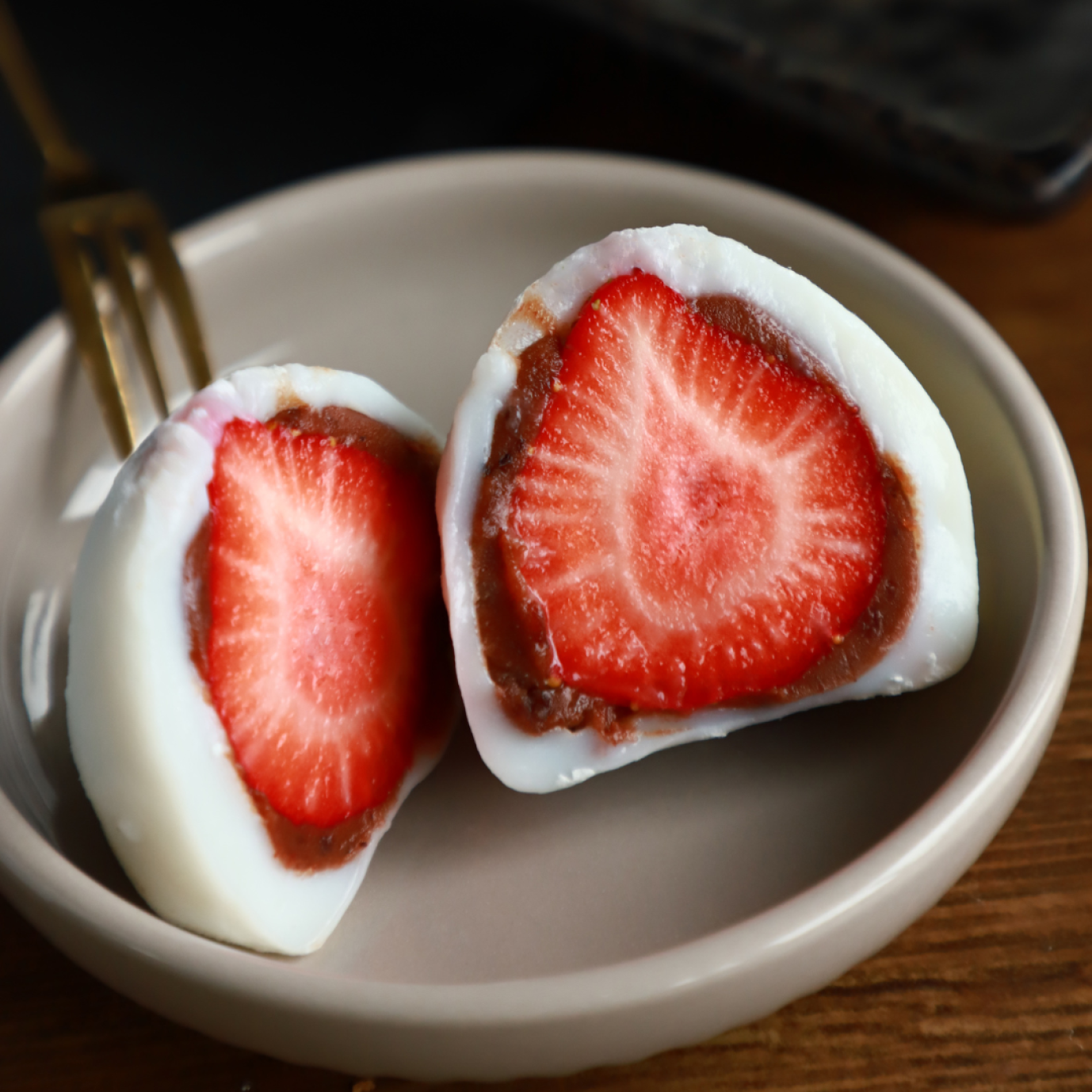 A close-up of two halves of strawberry daifuku - Japanese mochi filled with a whole strawberry and red bean paste - on a small beige plate with a fork in the background.