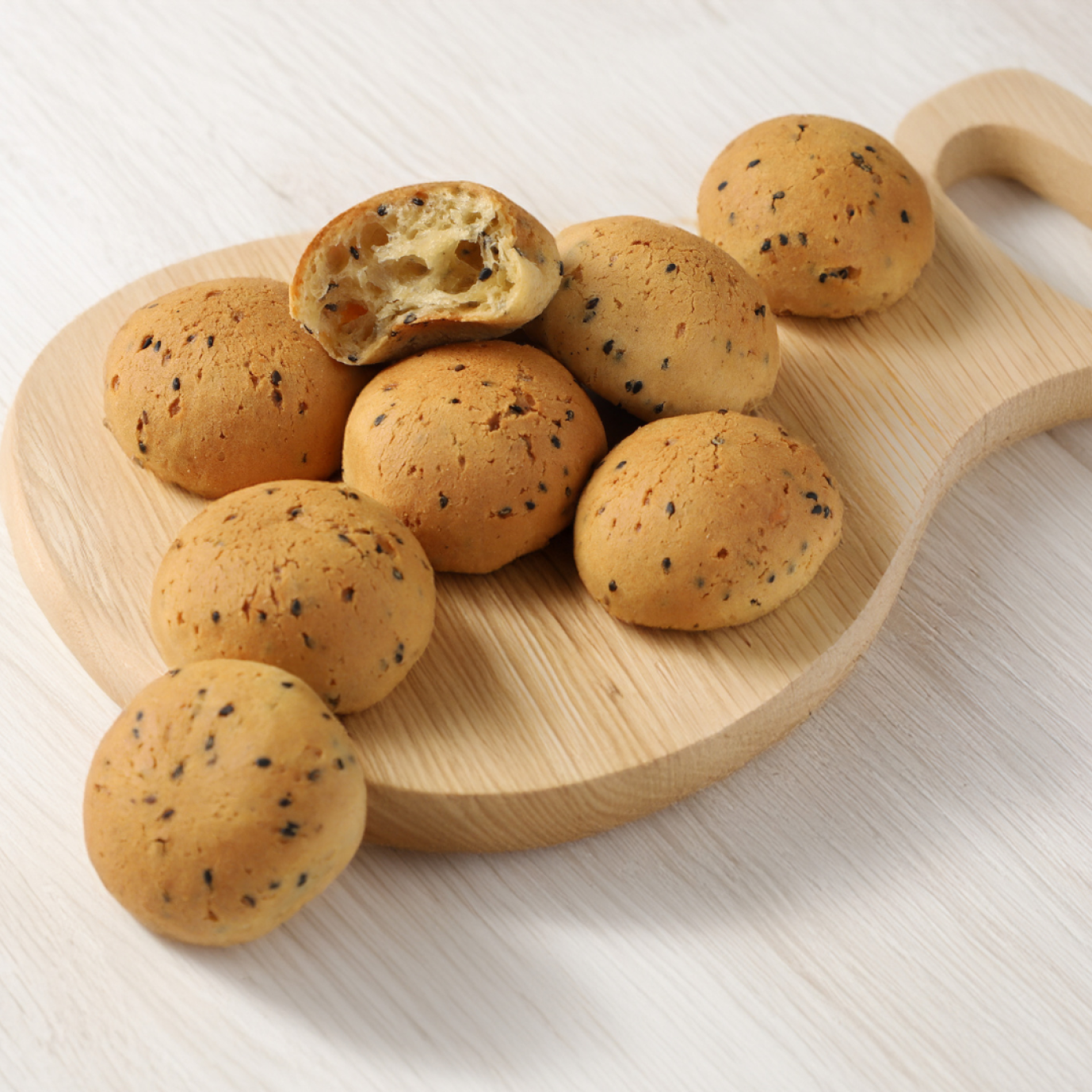 Round, golden-brown buns with black sesame seeds on a wooden serving board. One roll is broken open to reveal the soft, airy interior. The whole thing lies on a light-colored base.
