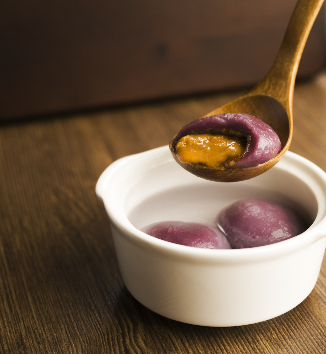 A wooden spoon holds a purple sticky rice ball with a black sesame filling over a white bowl containing more rice balls in clear liquid, set on a wooden table.